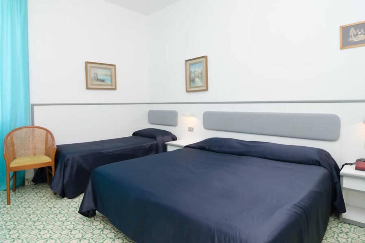 A hotel room with two beds, a chair, a desk, and two framed pictures on the wall.