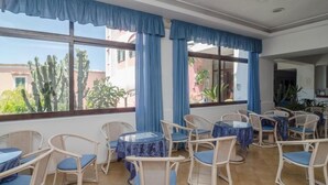 Una habitación con sillas y mesas de mimbre, ventanas amplias con cortinas azules, y vistas de cactus y edificios al exterior.