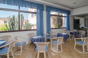 A room with wicker chairs and tables, large windows with blue curtains, and a view of cacti and buildings outside.