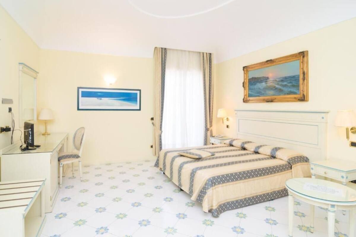 A hotel room with a bed, a desk, a chair, a small table, and a painting on the wall.