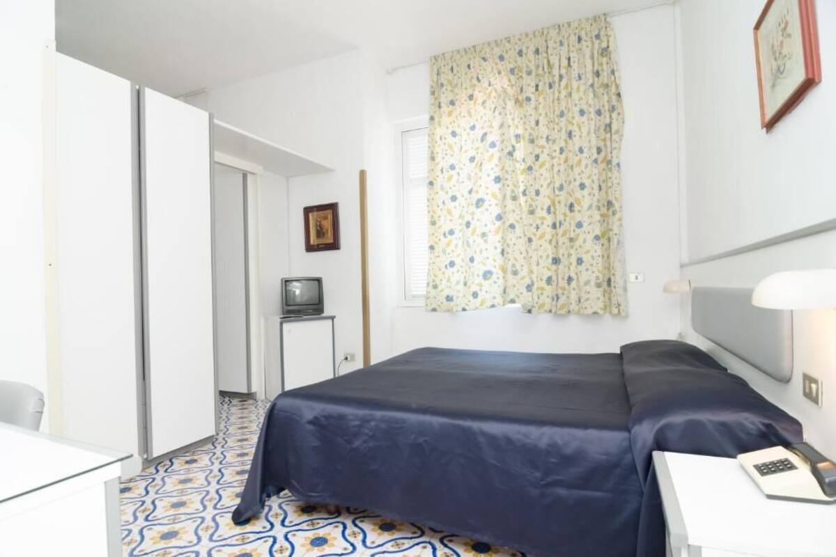 A hotel room with a bed, a desk with a lamp, a television, and a patterned floor.