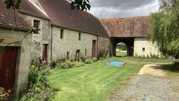 Un bâtiment rustique en pierre, doté d’un toit en pente, d’un chemin de gravier et d’une zone herbeuse parsemée de fleurs.