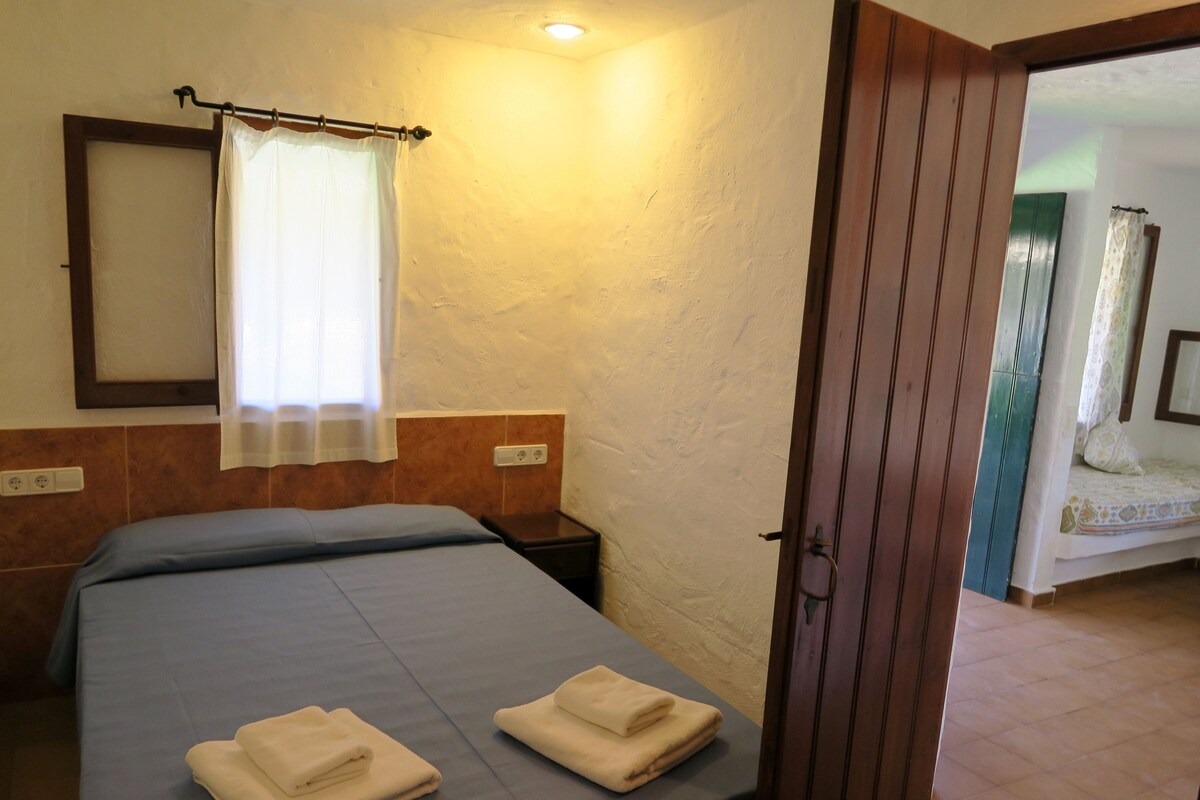 A bedroom with a bed, a nightstand, a wall-mounted light, and a door leading to another room.