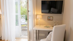 Une chambre d’hôtel avec un téléviseur fixé au mur, une table de chevet avec une lampe, une chaise et une vue sur le jardin par une porte ouverte.