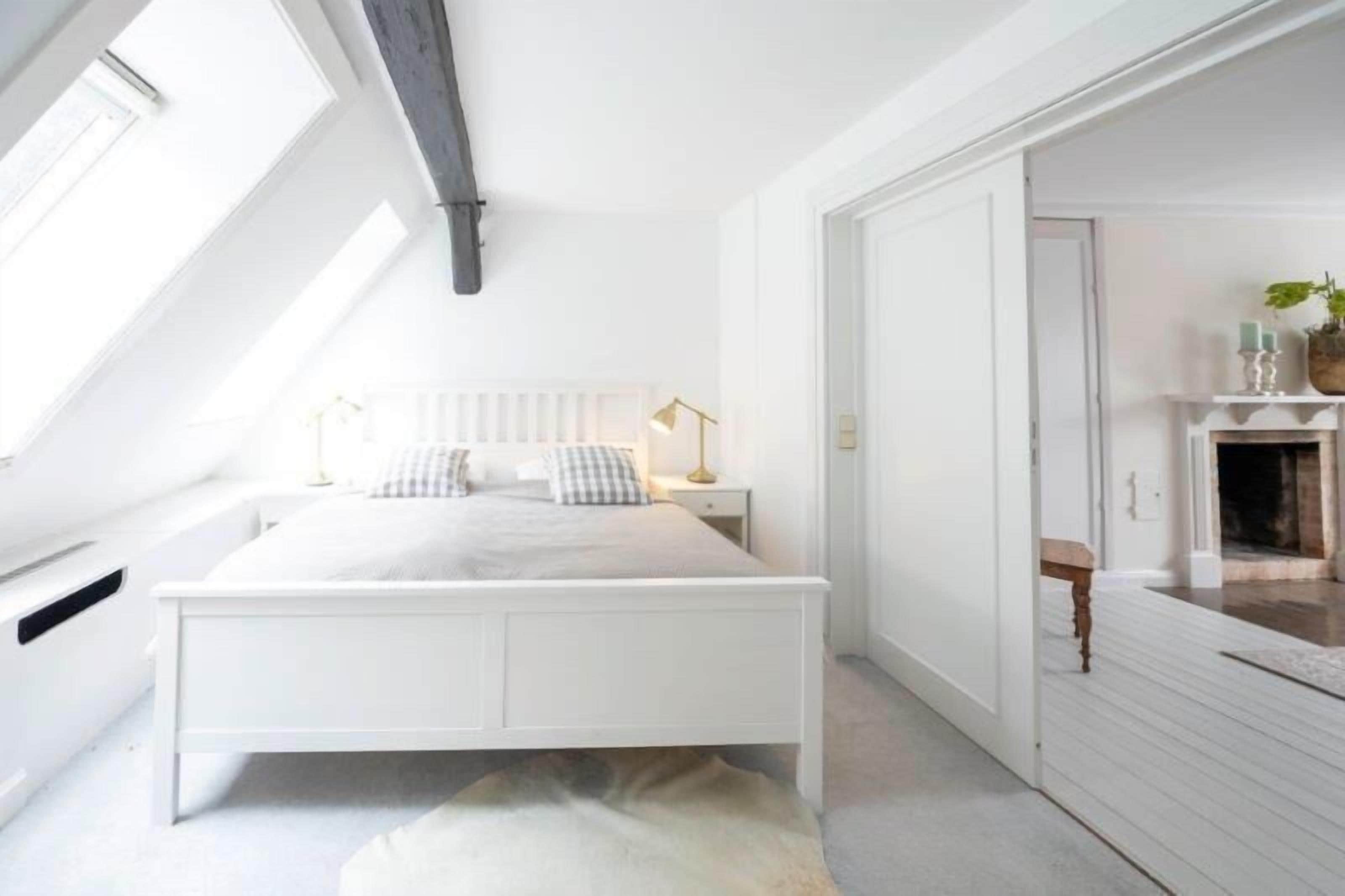 A bedroom with a white bed, a nightstand, a lamp, a radiator, and a fireplace.