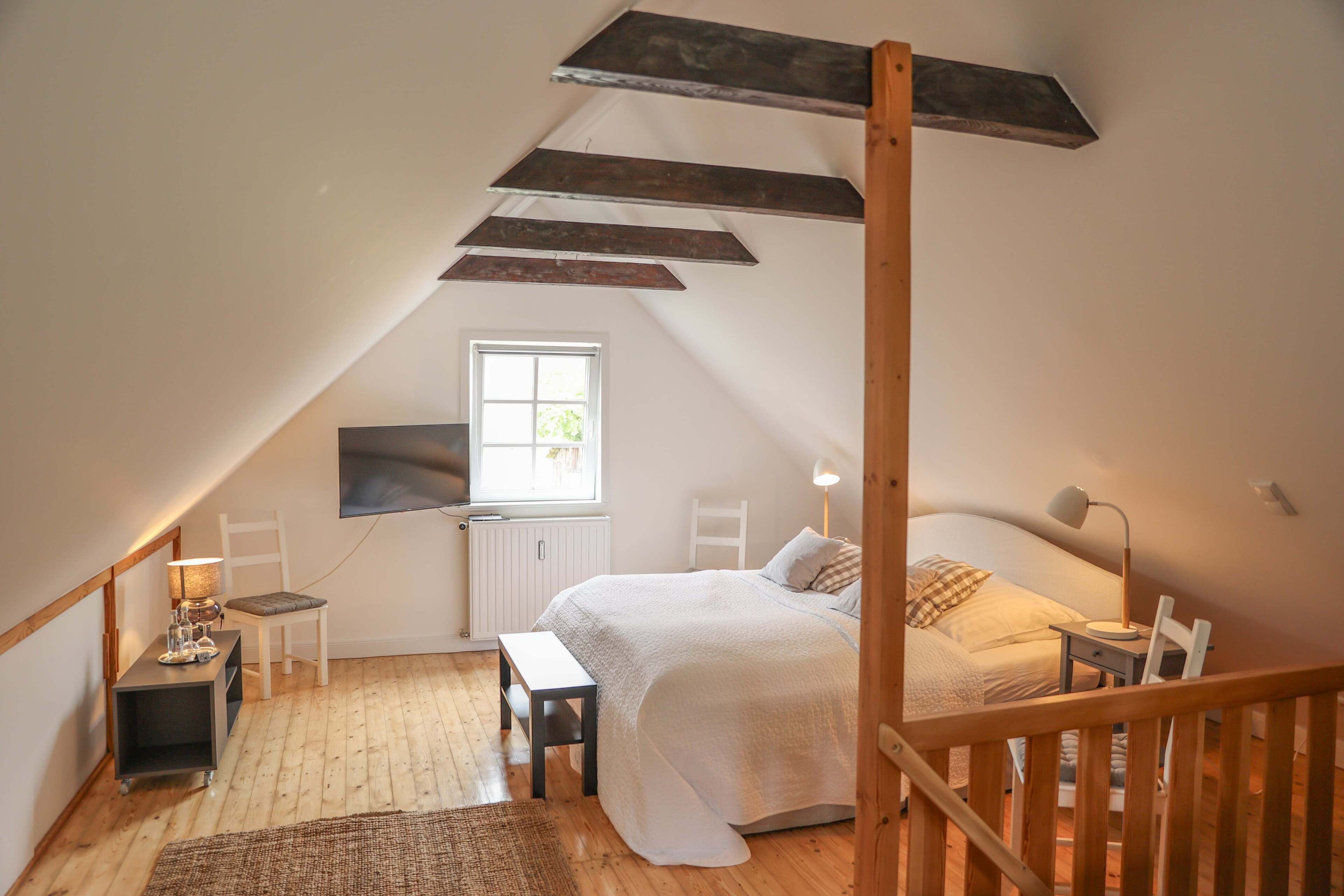 Un dormitorio acogedor en el ático, con techo inclinado, una cama grande, un escritorio, una silla y una ventana que proporciona luz natural.