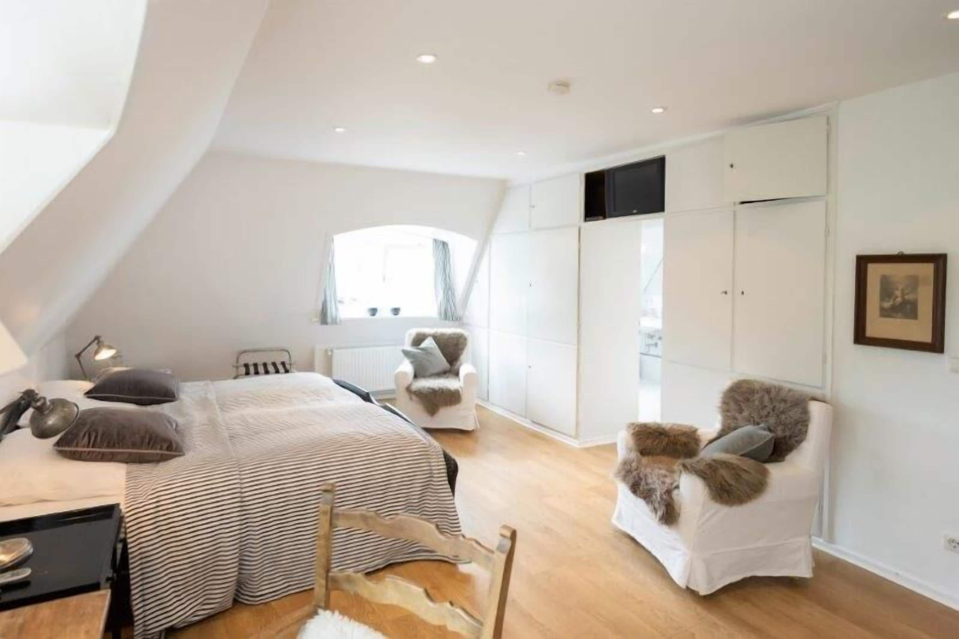 A bedroom with a bed, two chairs, a desk, a radiator, and a wardrobe.