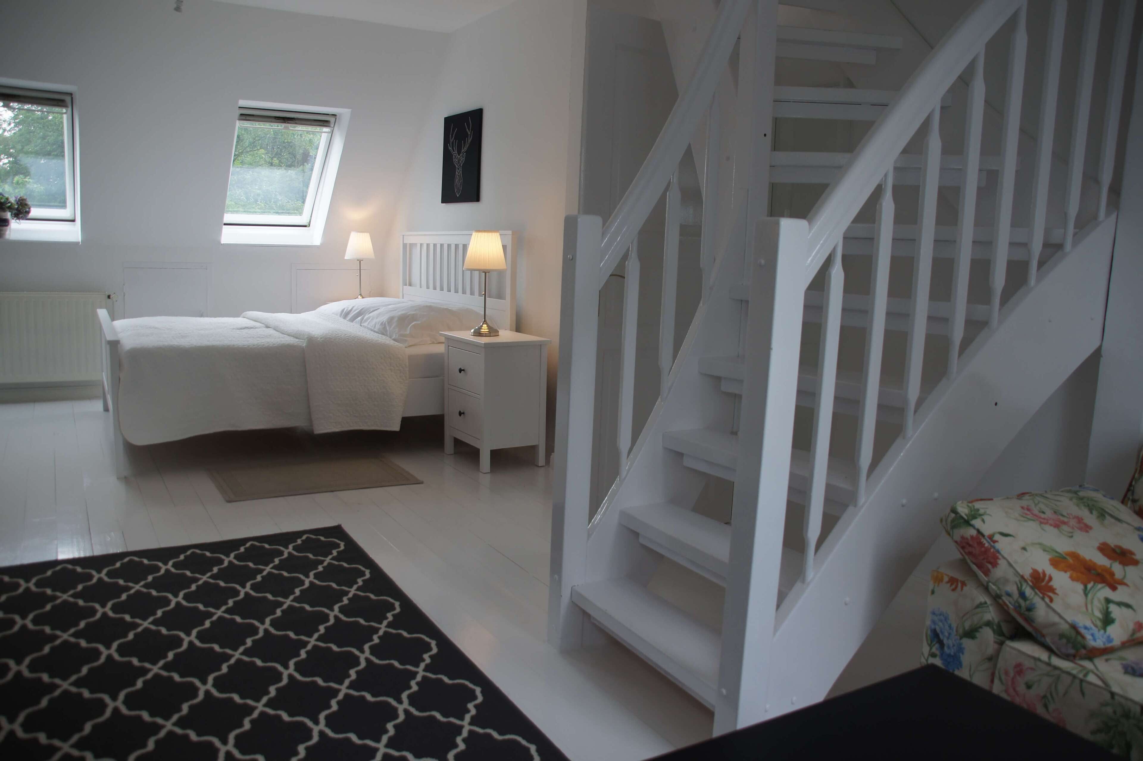 A bedroom with a bed, bedside tables, a lamp, a window, a radiator, and a staircase.