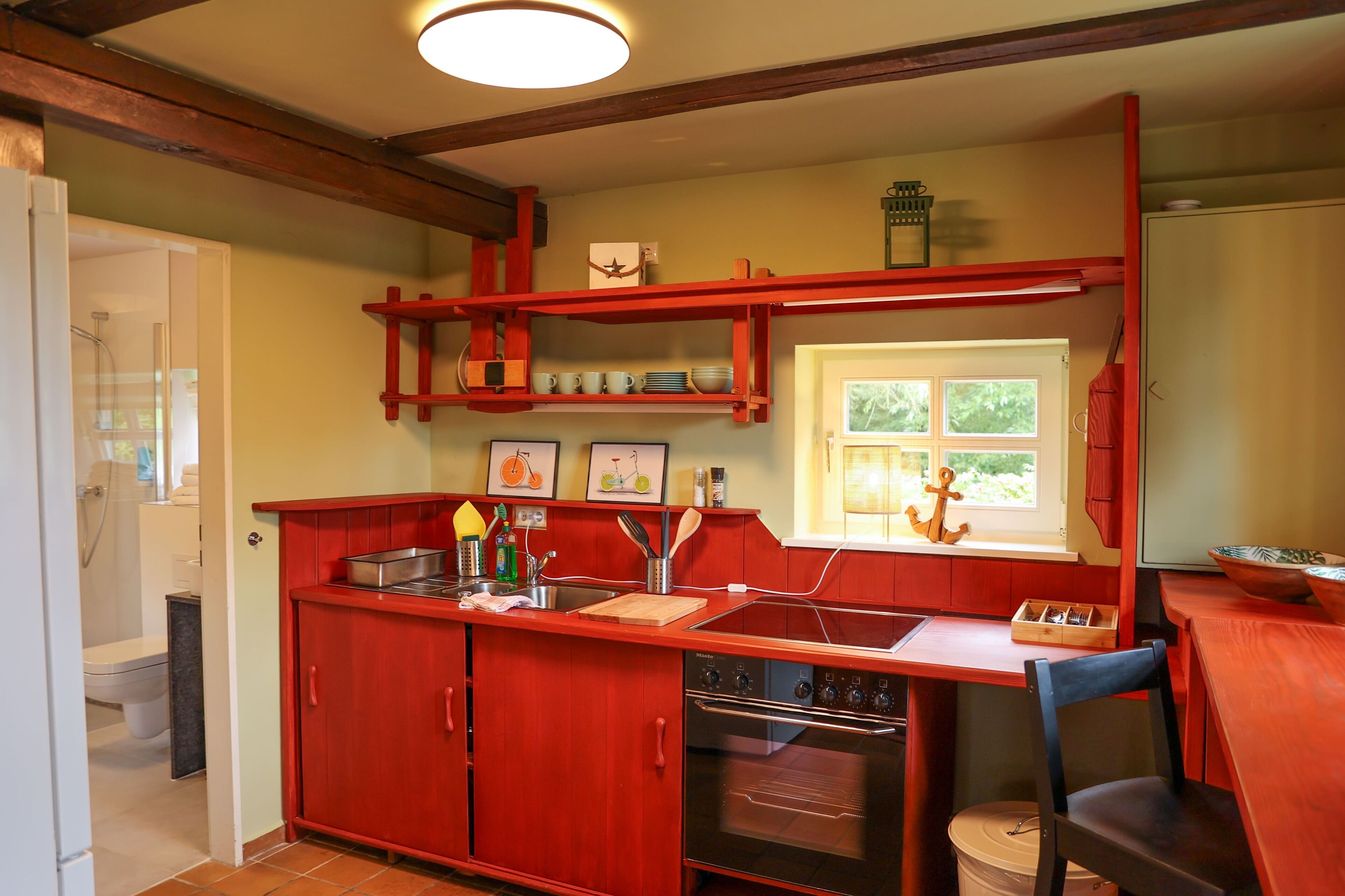 A compact kitchen with red cabinetry, a built-in oven, and a window with a view of greenery.