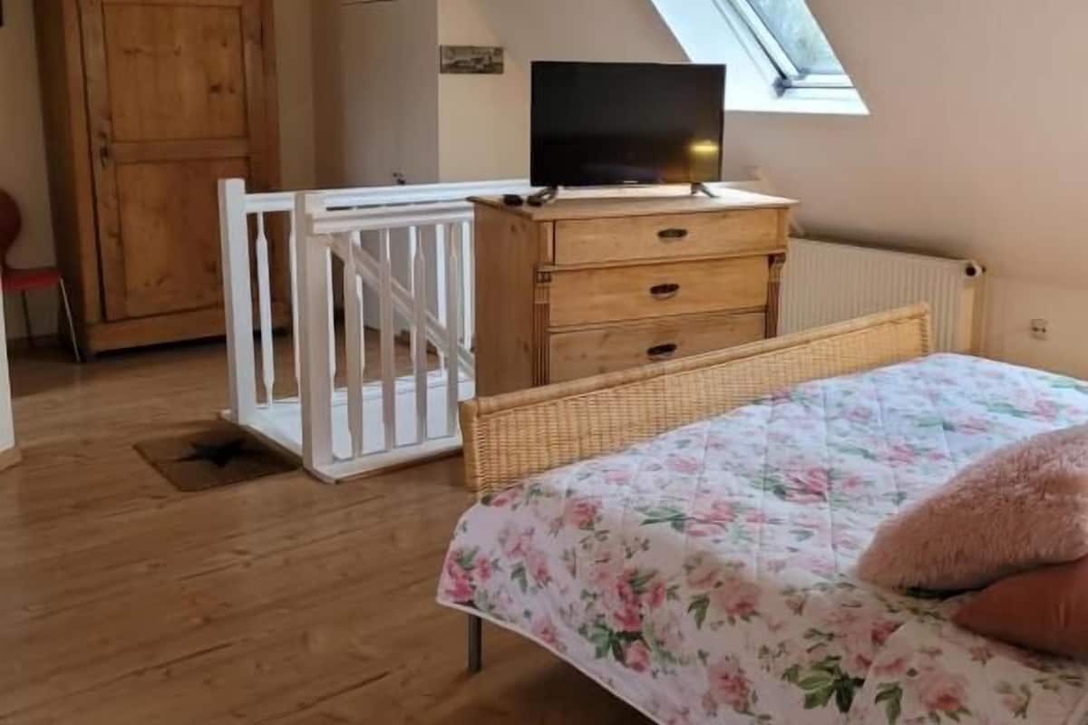 A bedroom with a sloped ceiling, a wooden wardrobe, a television on a dresser, a bed with floral bedding, and a skylight.