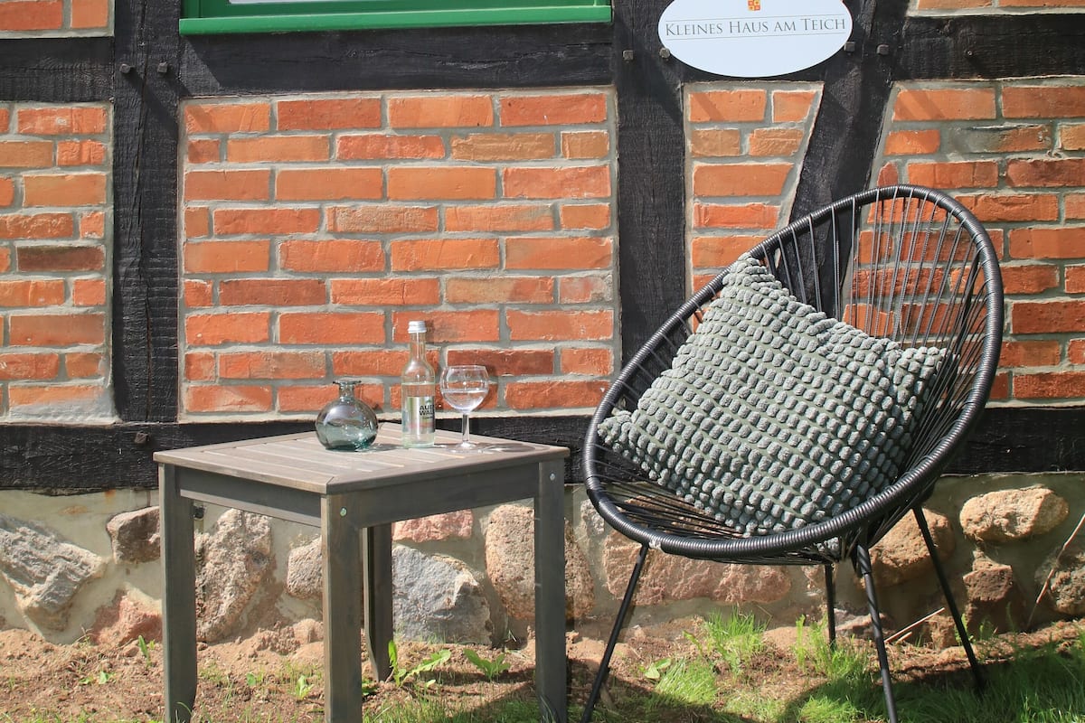 A table with a bottle and glass, a wicker chair, and a sign reading "Kleines Haus Am Teich" against a brick wall with a green window frame.