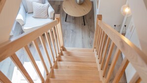 Un escalier en bois mène à une pièce où se trouvent un canapé blanc, une table ronde et un luminaire suspendu.