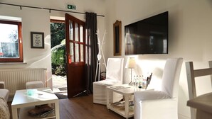 Une chambre d’hôtel avec un canapé blanc, une petite table avec une lampe, un téléviseur fixé au mur et une fenêtre avec des rideaux.