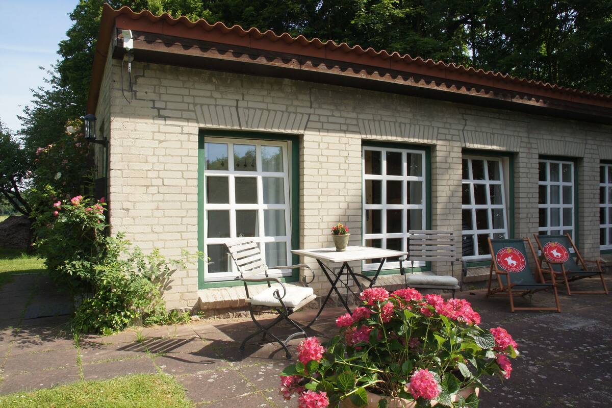 A building with a brick facade, large windows, outdoor seating, and a garden with pink flowers.