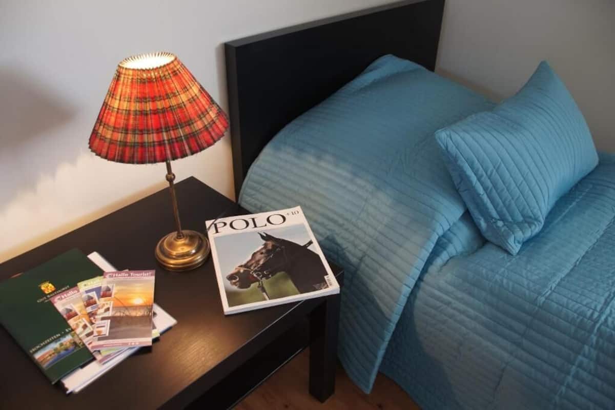 A neatly made bed with blue bedding, a magazine titled "POLO" on a bedside table, and a plaid lamp.