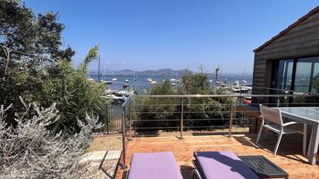 Une terrasse en bois avec deux fauteuils de jardin violets, une table en verre et une vue sur les bateaux qui naviguent.