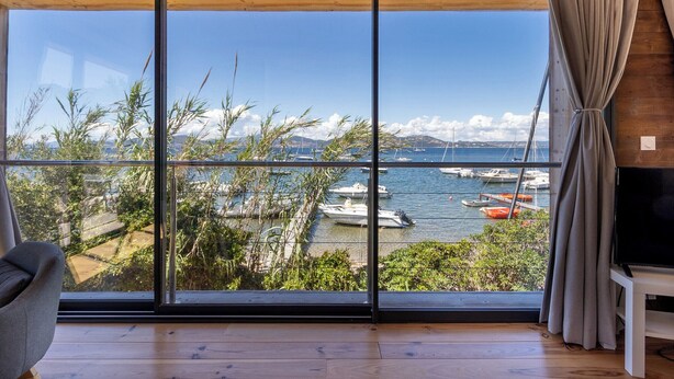 Habitación con ventanas amplias que ofrecen vistas a un puerto deportivo con barcos y montañas.