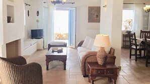 Una sala de estar con sofá, mesa de centro y lámpara. Se ve un televisor y un comedor al fondo.