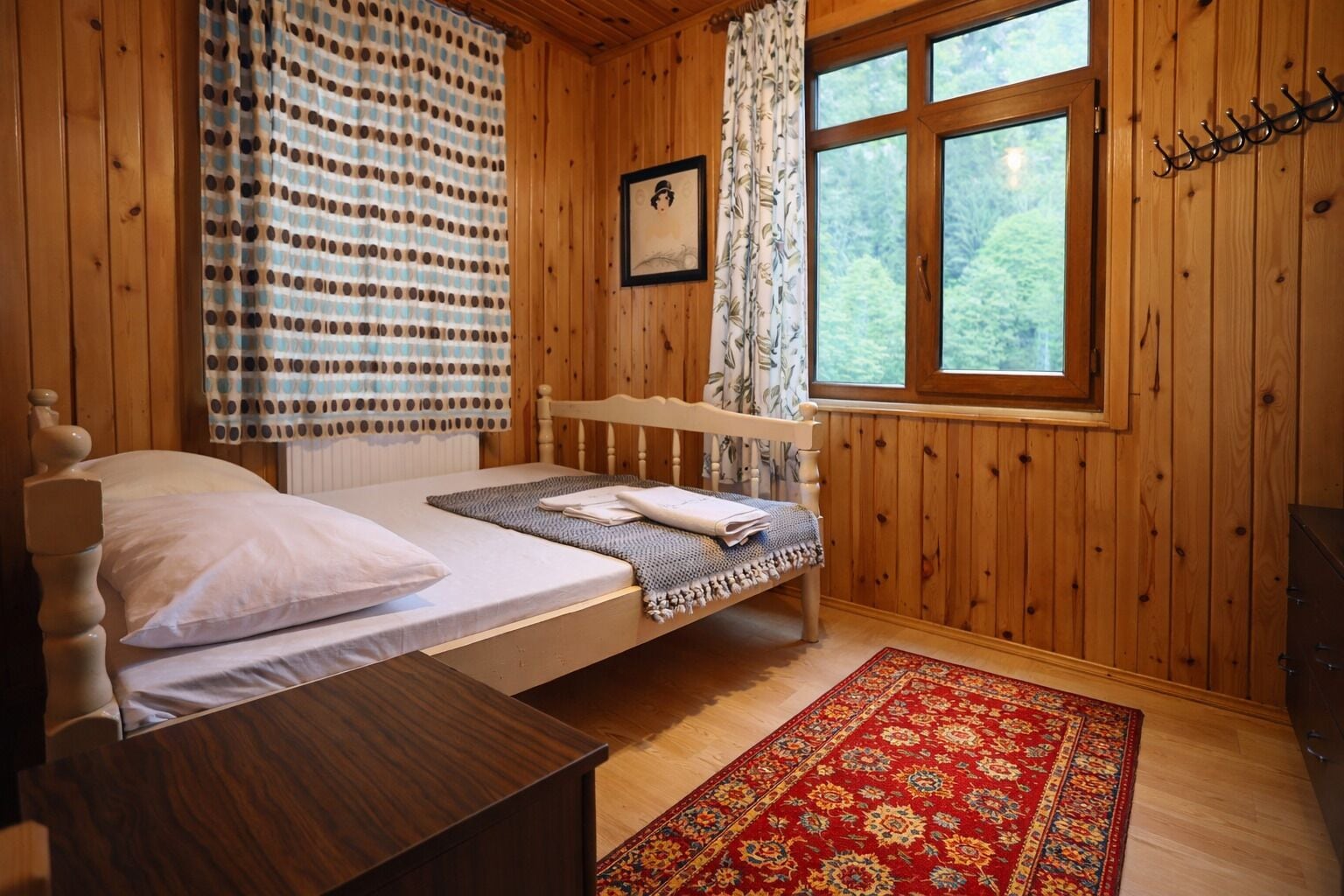A wooden cabin bedroom with a bed, a wooden headboard, a window with a view of trees, a patterned curtain, and a red rug.