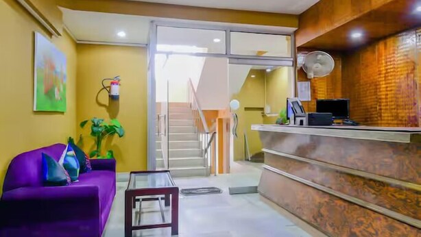 Un espace d’accueil avec un canapé violet, une table en verre, un comptoir d’accueil en bois, un escalier et un ventilateur fixé au mur.