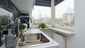 Une cuisine moderne avec un évier en acier inoxydable, une bouilloire et une plante en pot près d’une fenêtre donnant sur un paysage urbain.
