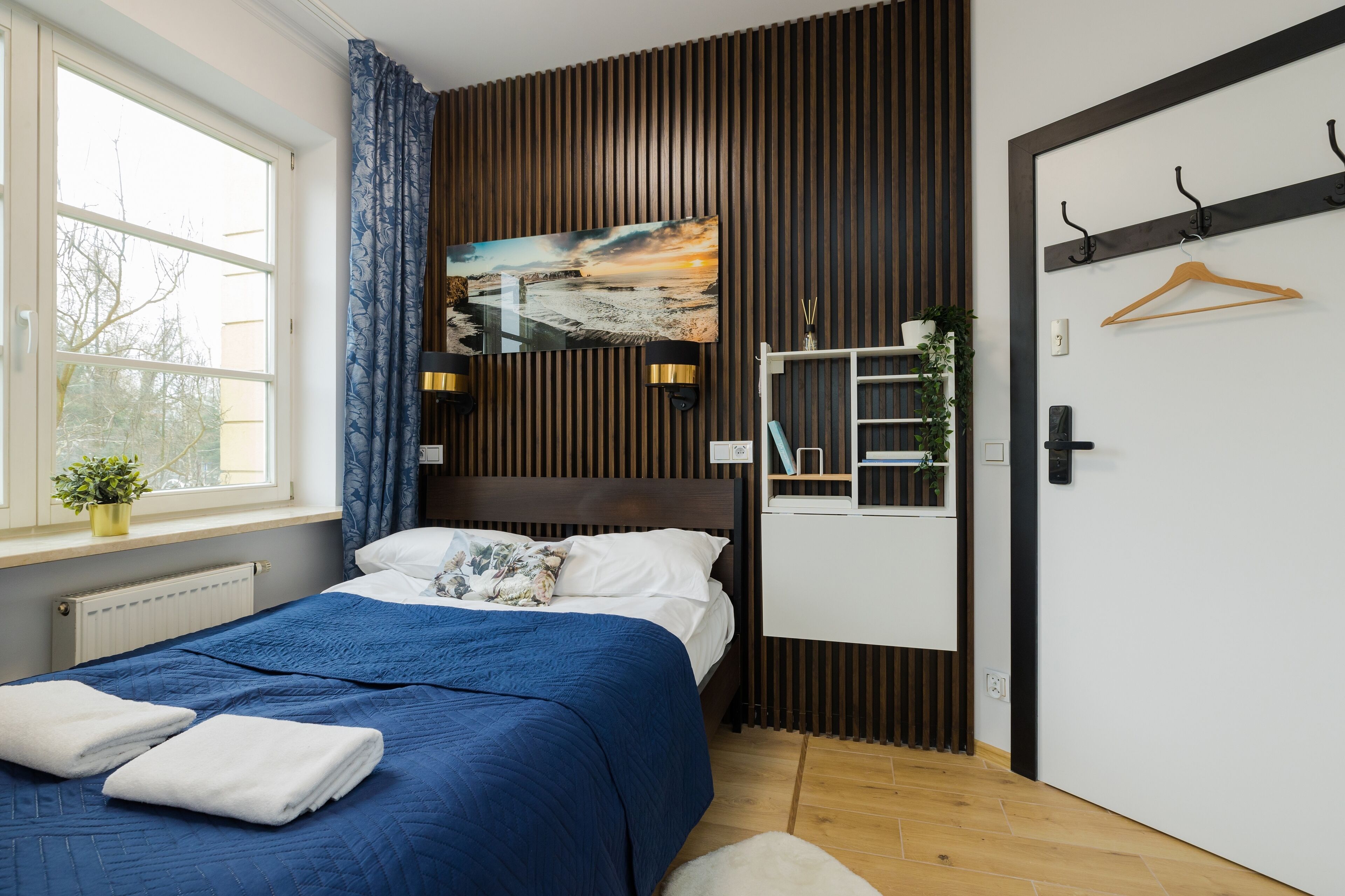 Une chambre à coucher avec un lit, une fenêtre avec des rideaux, un téléviseur fixé au mur, un porte-manteau et une porte.