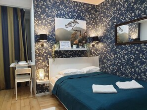 A bedroom with a bed, bedside lamps, a small table with a chair, and a wall-mounted shelf with plants.