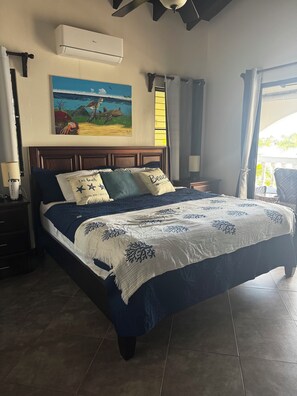 A bedroom with a bed, a painting, an air conditioning unit, a nightstand, a lamp, and a view of the outdoors.