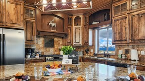 A kitchen with wooden cabinets, a stainless steel refrigerator, granite countertops, and a large island with breakfast items. - Spacious Cabin with Hot Tub, Fire Pit, Game Room (Island Park,)