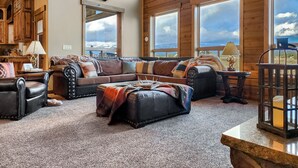 A spacious living room with a large wooden ceiling, a mounted deer head, a leather sofa set, and a view of the outdoors through large windows. - Spacious Cabin with Hot Tub, Fire Pit, Game Room (Island Park,)