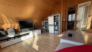 Un soggiorno moderno con pareti in legno, una TV a schermo piatto, una libreria e un cuscino rosso su un poggiapiedi bianco.