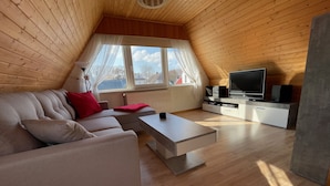 Un soggiorno moderno in stile loft con soffitto in legno, un'ampia finestra con tende, un divano beige con cuscini rossi, un tavolino da caffè, una TV e un altoparlante.
