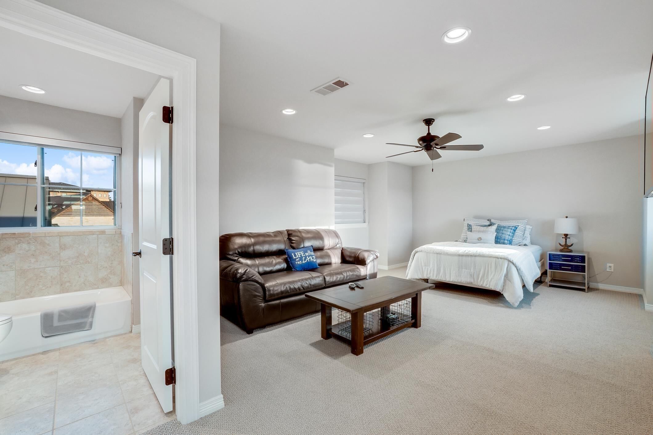 A bedroom with a bed, a brown leather sofa, a coffee table, a ceiling fan, and a view of a balcony.