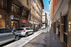 Uma rua estreita, ladeada por edifícios altos, carros estacionados e uma fileira de bicicletas.