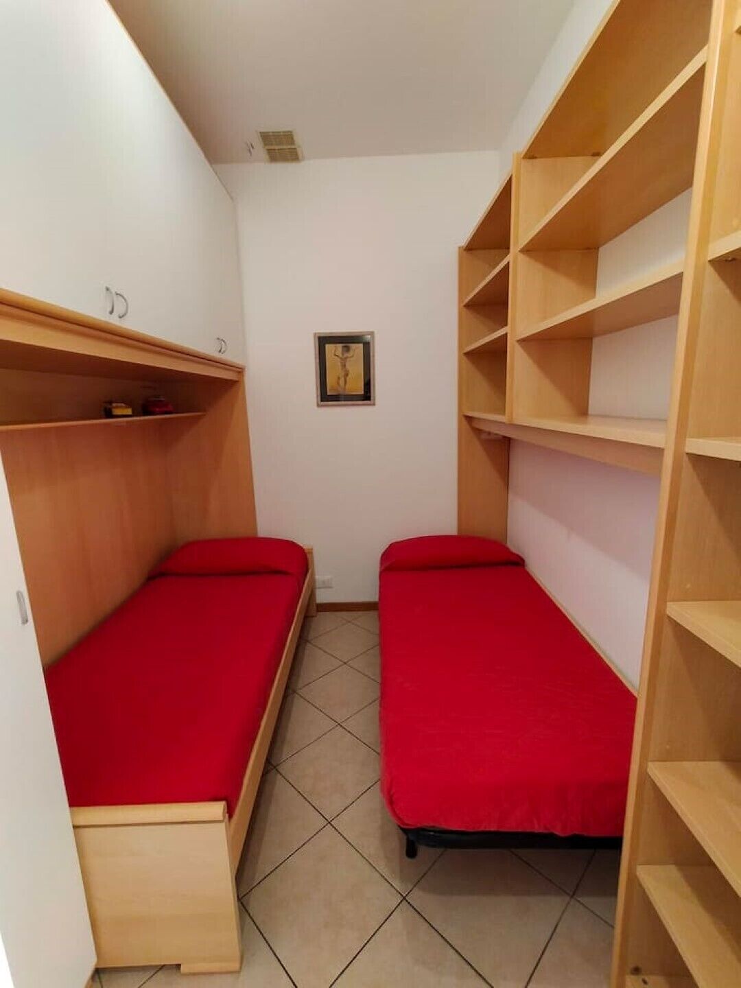 A small, narrow room with two single beds, wooden shelves, and a framed picture on the wall.