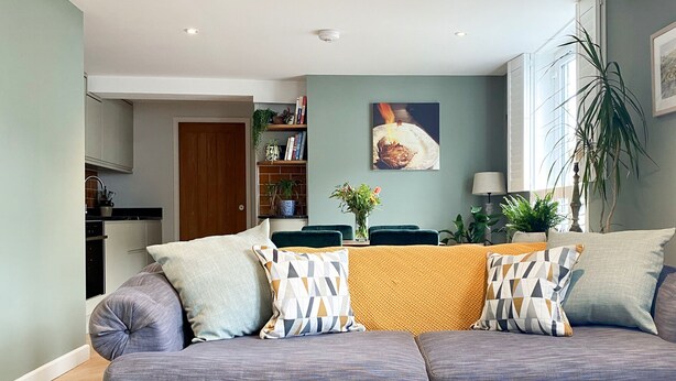 A modern living room with a grey sofa, patterned cushions, a round coffee table with a vase, and a dining area in the background. - The Old Stables - Luxury Dartmouth town centre apartment w/ parking permit (Dartmouth)