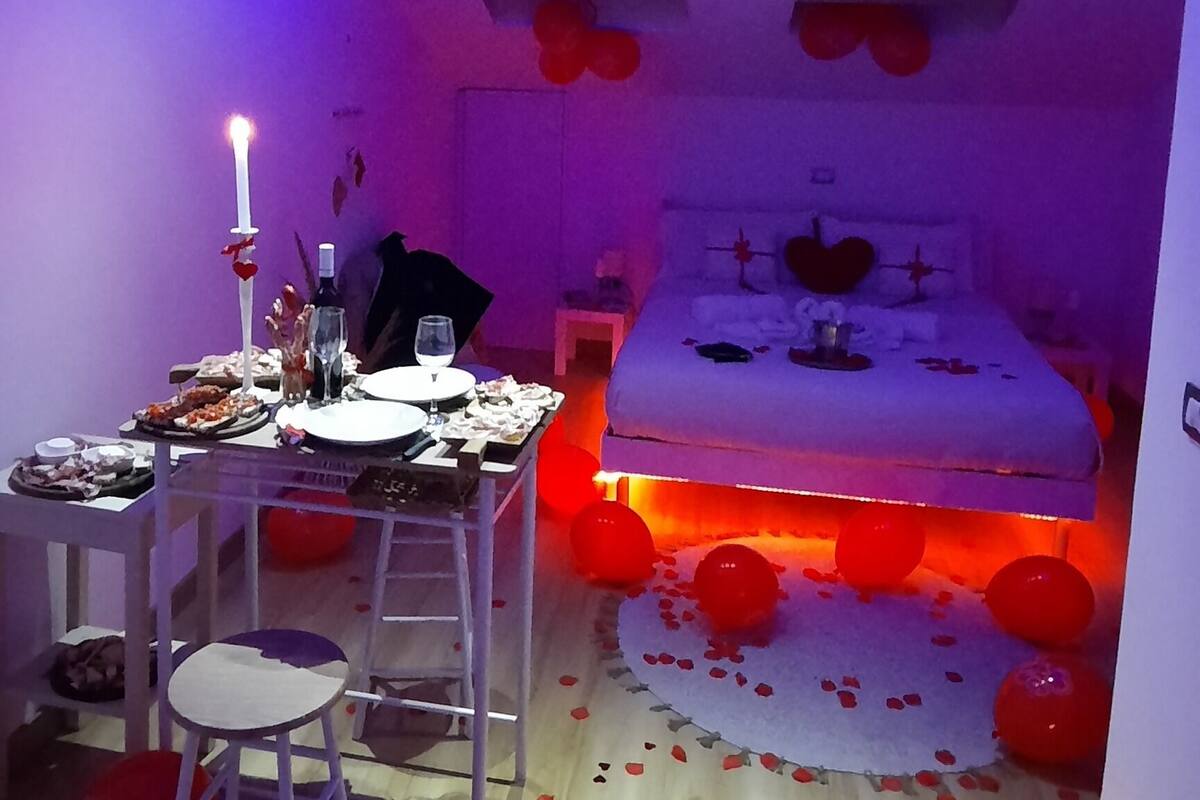 A bedroom with a bed, a bedside table, and a chair. There are red balloons and rose petals on the floor.