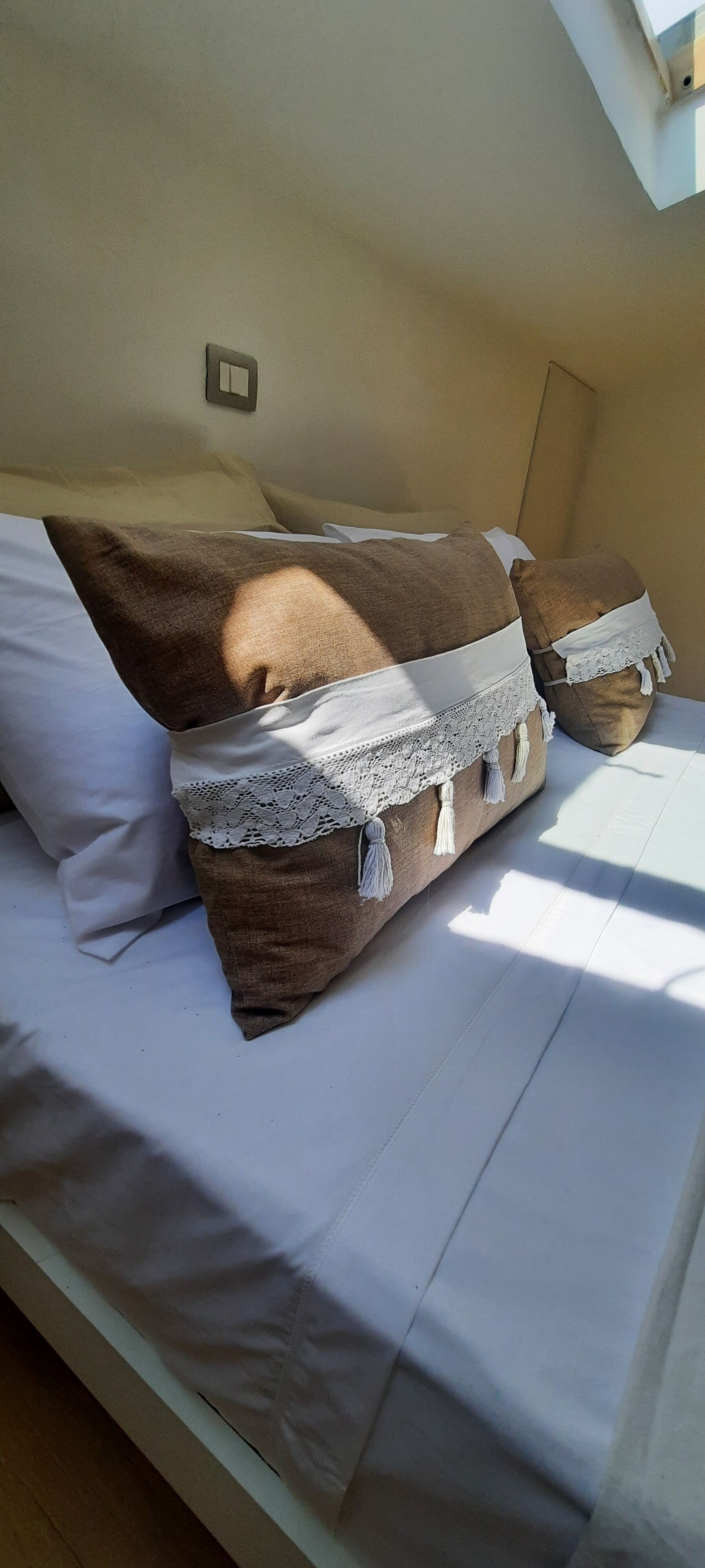 Una cama con sábanas blancas y almohadones decorativos, una mesita de noche pequeña y un interruptor de luz en la pared.