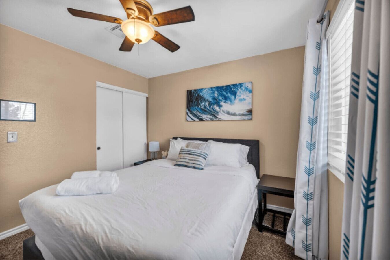 A hotel room with a bed, a ceiling fan, a painting, and a window with curtains.