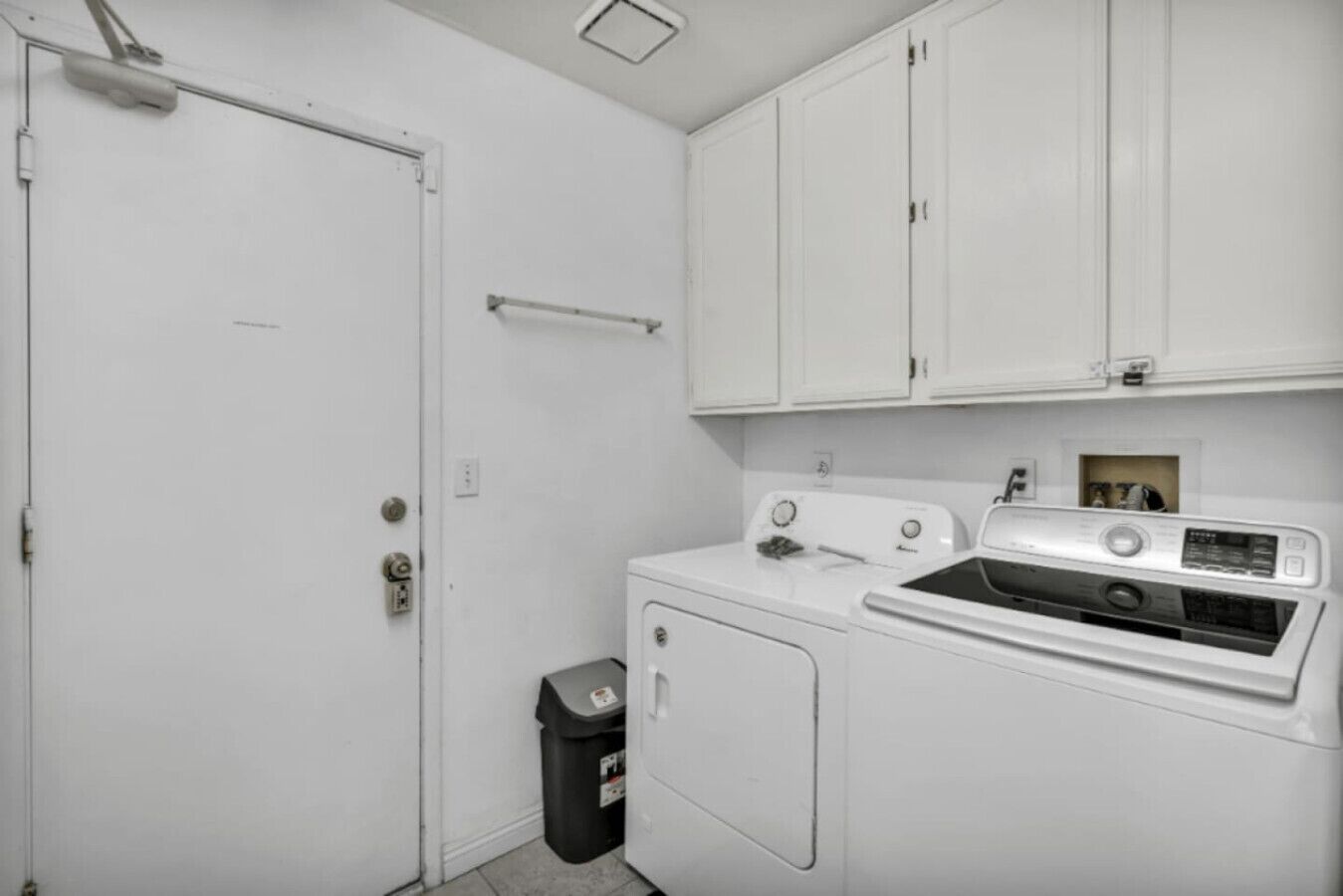 A laundry room with a washer and dryer, a trash can, and white cabinets.