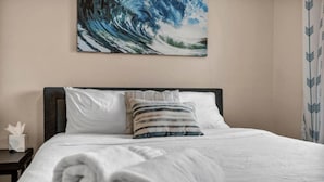 A neatly made bed with white linens and a rolled towel, a striped pillow, and a painting of a wave on the wall above.