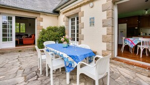 Een terras van natuursteen met een tafel en stoelen, een deur naar een keuken, en een kleine tuin.
