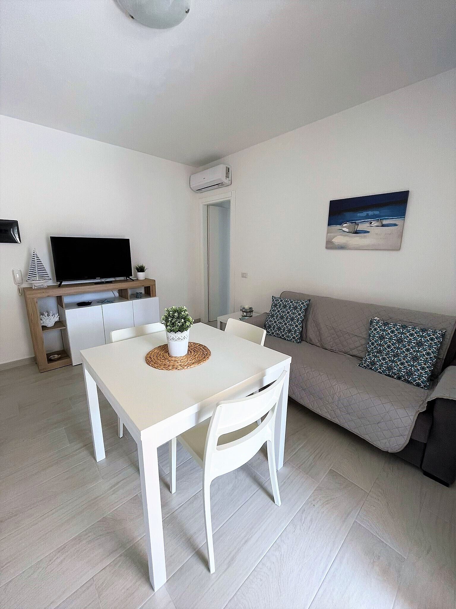Una sala de estar moderna con una mesa de comedor blanca, sillas blancas, un sofá gris con cojines estampados, un televisor de pantalla plana sobre un mueble de madera y un cuadro en la pared.