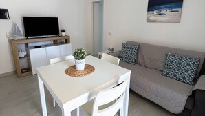 Un salon moderne avec une table à manger blanche, des chaises blanches, un canapé gris agrémenté de coussins à motifs, un téléviseur à écran plat posé sur un meuble en bois et un tableau accroché au mur.