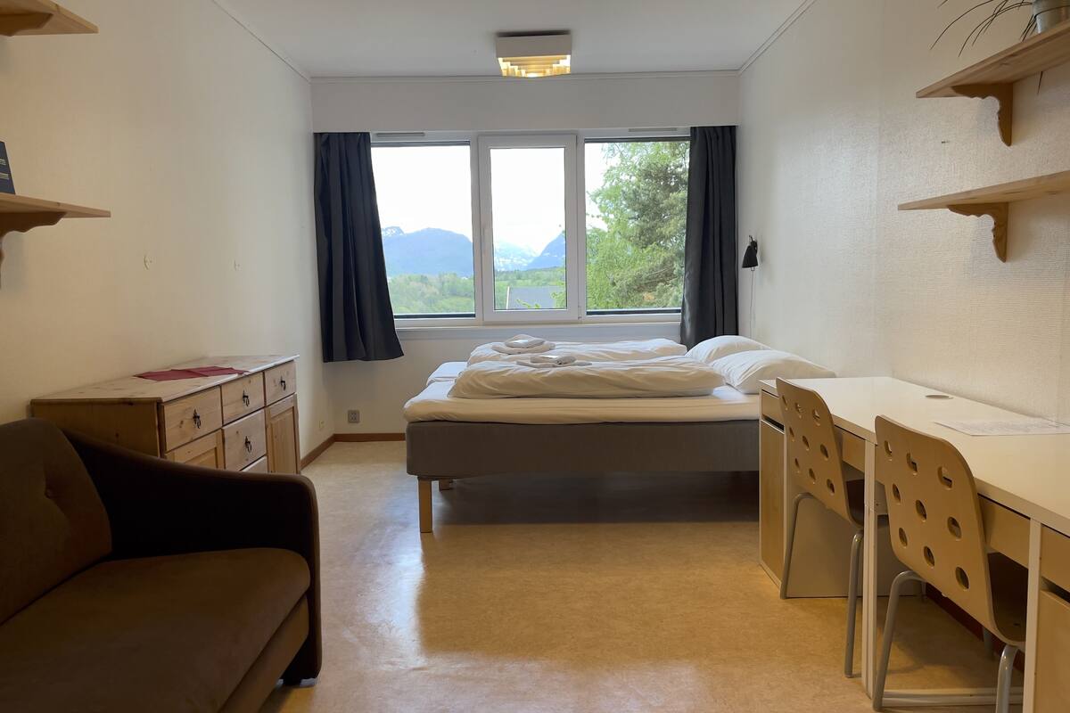 A bedroom with a bed, a desk, a chair, a dresser, and a window with a view of greenery and mountains.