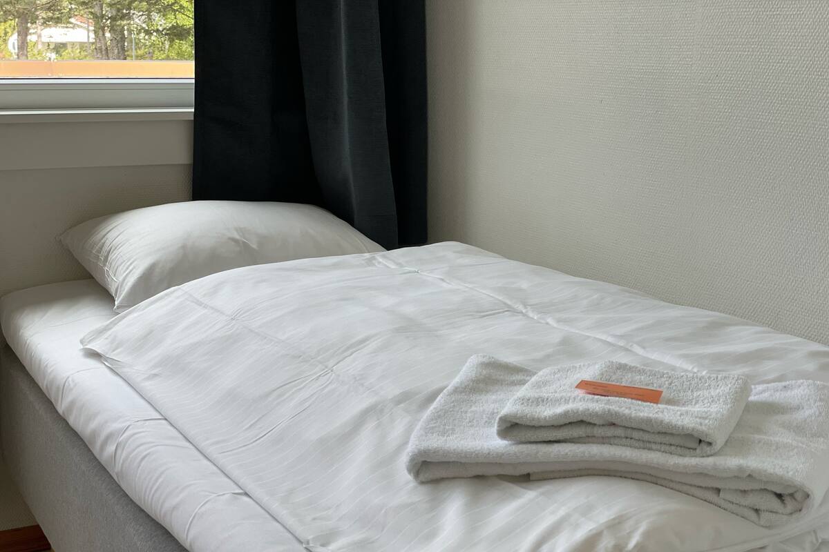 A neatly made bed with white linens, a folded towel on the side, and a window with curtains.