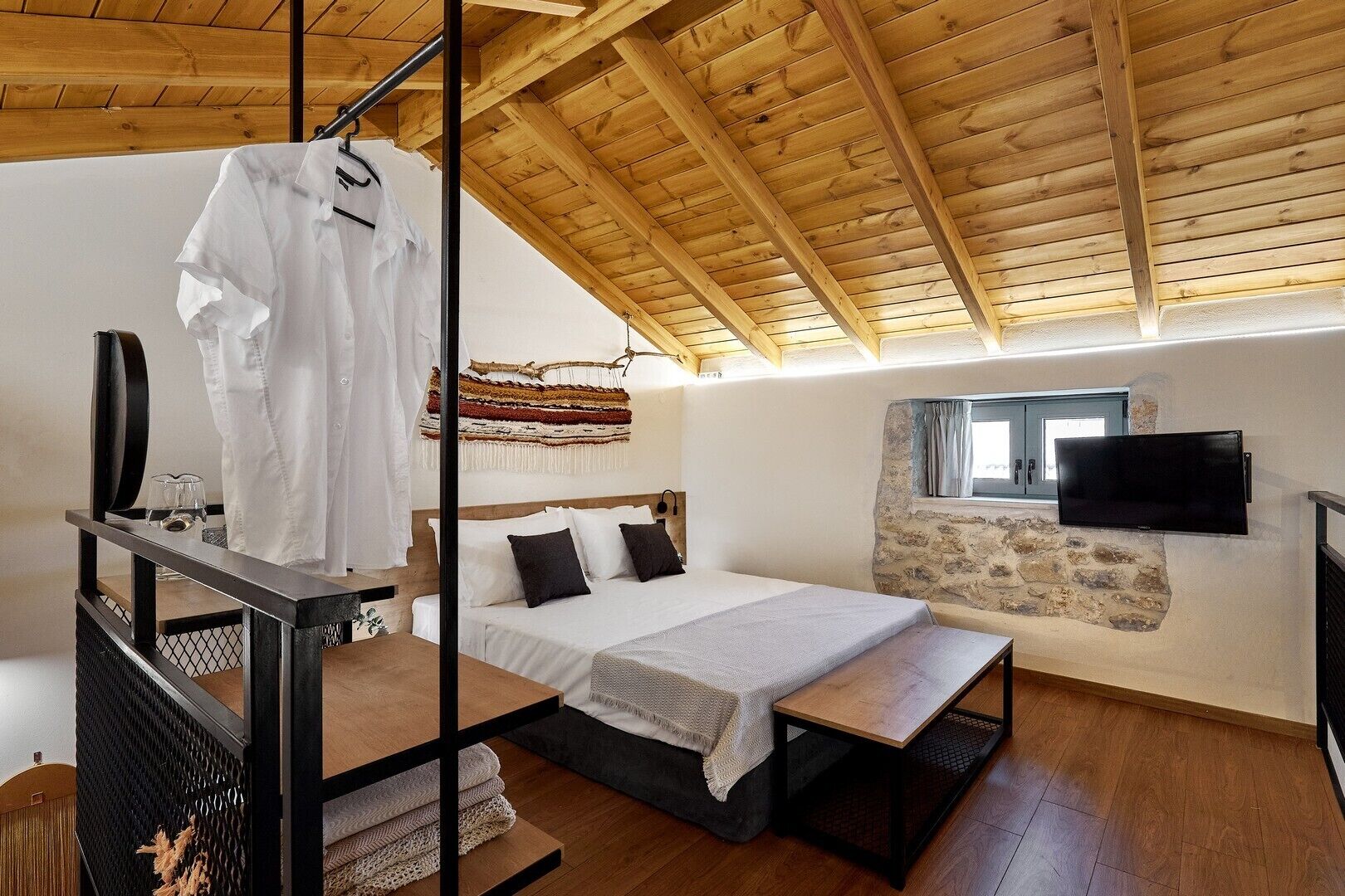 Una camera da letto con soffitto in legno, un letto con testiera, una TV a parete e una finestra con tende.