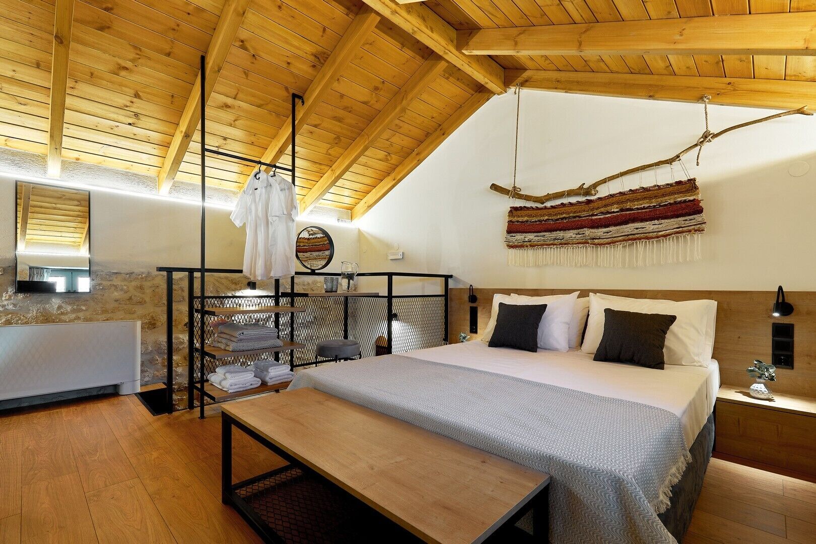 Un dormitorio acogedor con techo de madera, una cama con muchos almohadones, un banco y una decoración textil colgante.