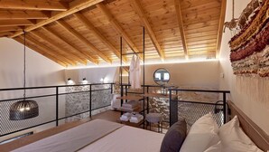 A cozy bedroom with a wooden ceiling, a bed with pillows, a small table with towels, and a hanging light fixture. - Vin Dimora Suites with shared pool - Vino Roze (Ano Archanes)