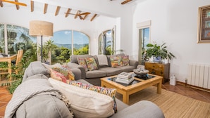 A spacious living room with a grey sofa, colorful pillows, a wooden coffee table, and a large window offering a view of greenery and mountains. - 3 Bedroom Family Villa with Pool (Javea / Xàbia)